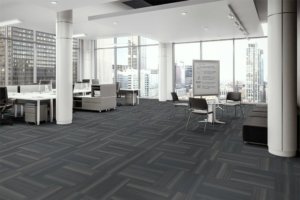 Fiction flooring in the open office. At one side are occupied work desks with attached credenzas. A portable white board is placed to the side of one round cafe table, with instructions written upon it. The full height windows look out to the city.